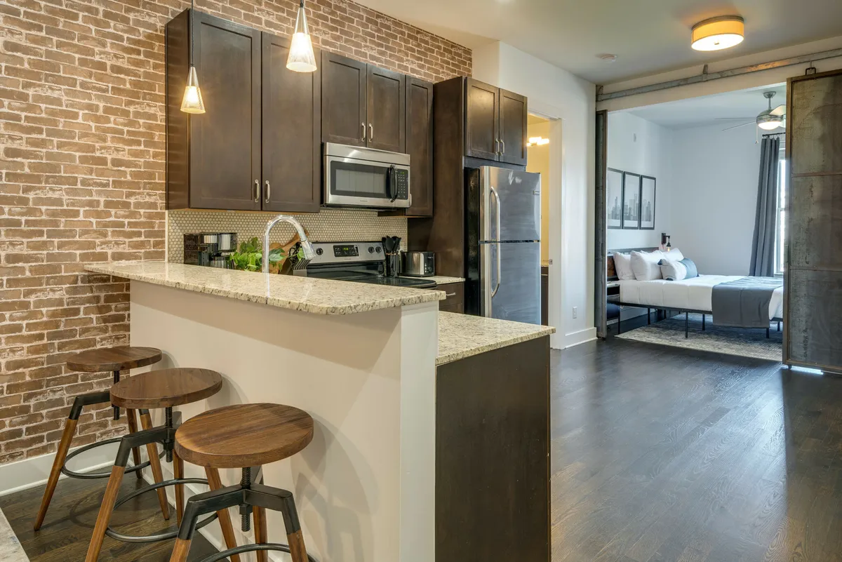 This image showcases a cozy, modern kitchen with sleek granite countertops, dark wood cabinets, exposed brick wall, and bar stools. Stainless steel appliances and decorative touches create an inviting atmosphere ideal for cooking, casual dining, or gathering with friends.