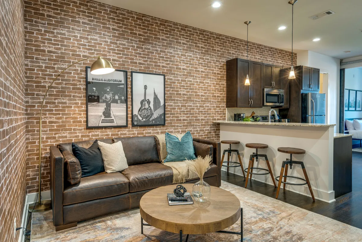The image showcases a stylish, open-concept living space with a rich leather sofa set against an exposed brick wall, complemented by tasteful artwork and a sleek kitchen bar area. The scene radiates warmth and creativity, inviting relaxation or socializing in a contemporary urban setting.