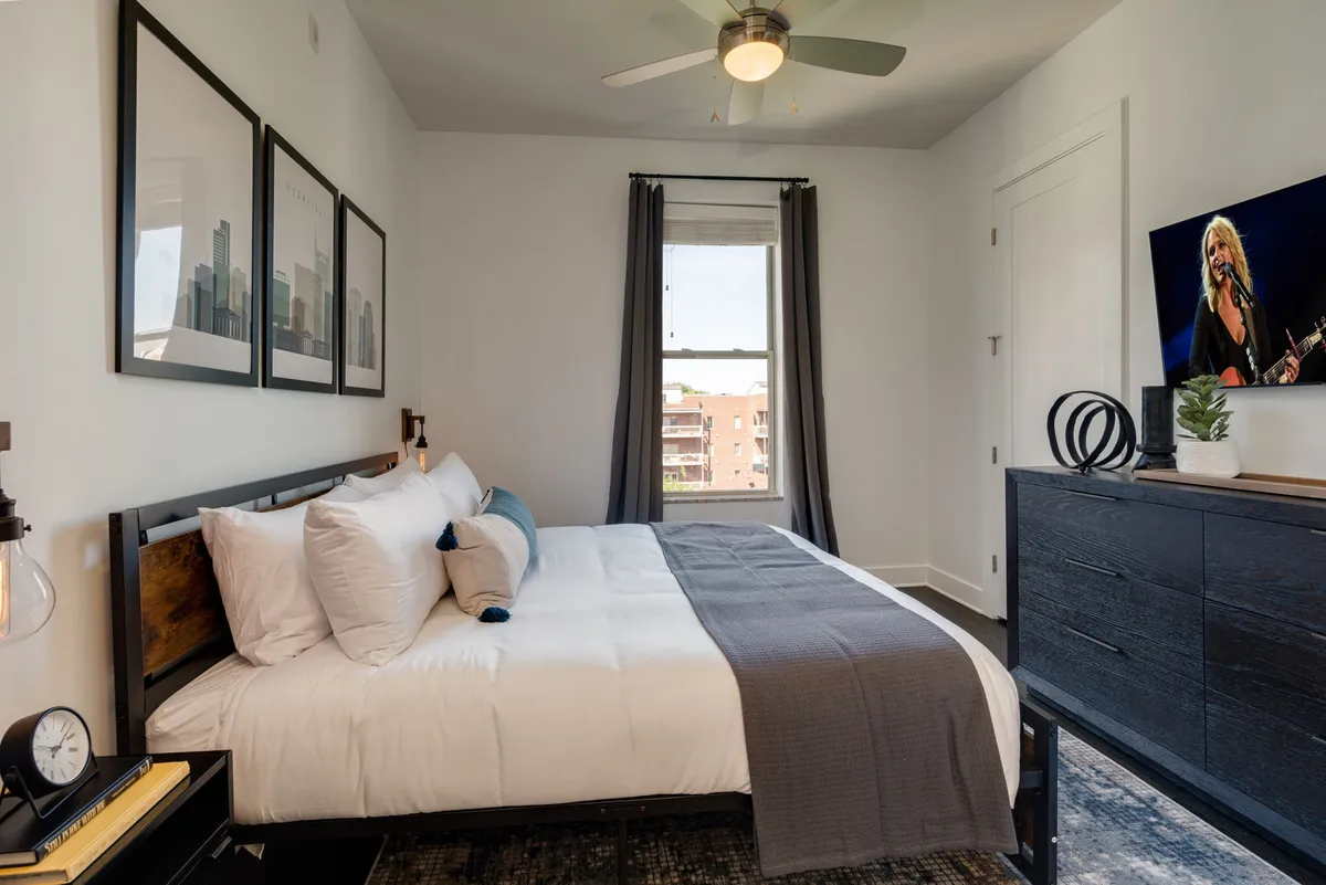 A modern bedroom with a comfortable bed, a window letting in natural light, a wall-mounted smart TV across from the bed, and tasteful artwork. The cozy setting highlights both entertainment and relaxation features.