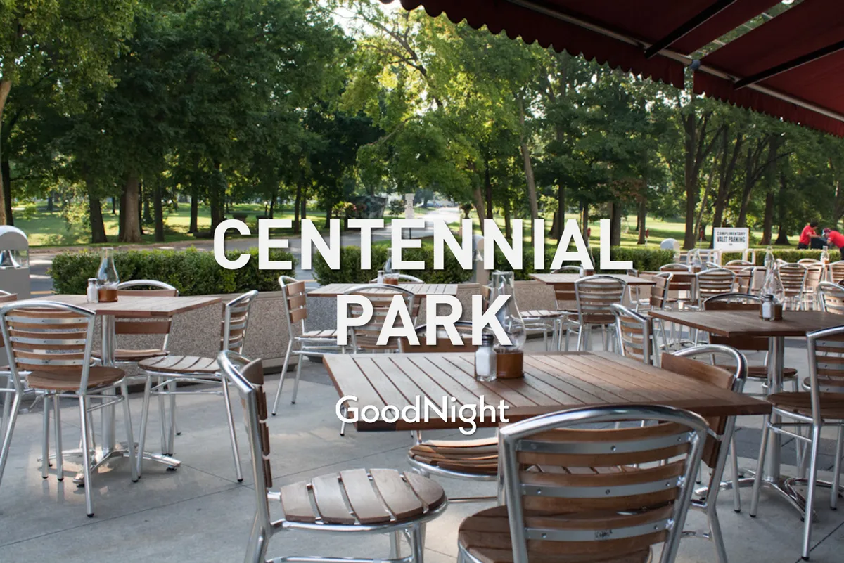 The image features outdoor seating at wooden tables surrounded by green foliage and a paved park pathway. The mood is welcoming and relaxed, suggesting the possibility of dining or socializing al fresco in a vibrant neighborhood setting.