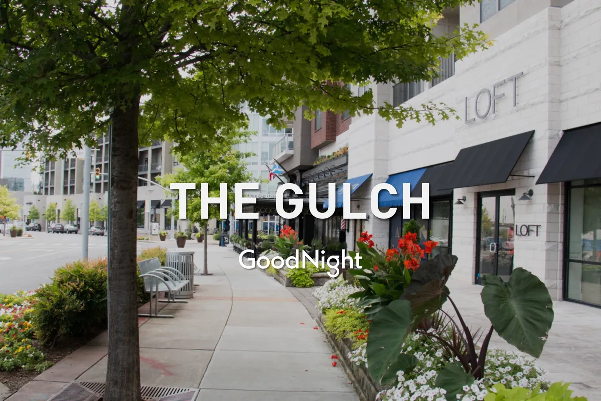 A welcoming streetscape with greenery, flower beds, benches, and storefronts. The atmosphere is friendly and open, with community-oriented energy perfect for family strolls and day-to-day conveniences.