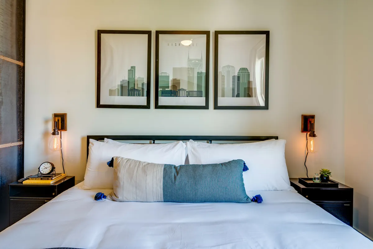 A calm and cozy bedroom featuring a neatly made bed with plush linens, framed artwork above the headboard, matching nightstands, and soft lighting fixtures. The atmosphere is serene and designed for relaxation, with an emphasis on comfort and tranquility.