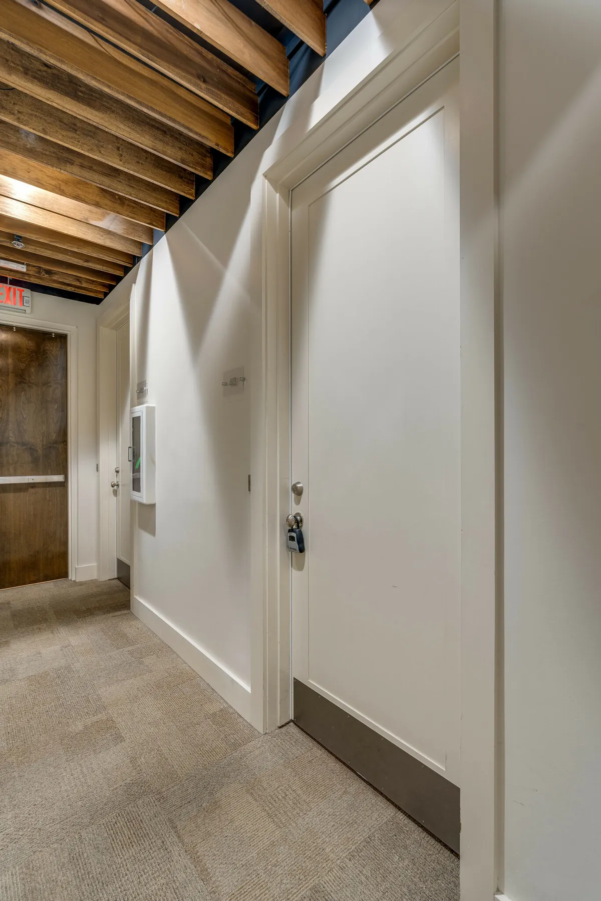 This image captures an interior view of an entryway area with wooden ceiling beams, crisp white doors, and a carpeted floor, all set in a calm and orderly mood. The scene suggests entering or exiting a private unit and hints at quiet moments of arrival.